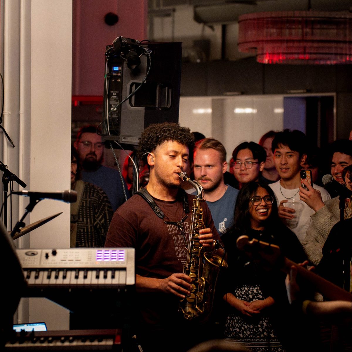 A saxophonist playing beside a keyboard to a standing crowd at an evening event at Den 1880