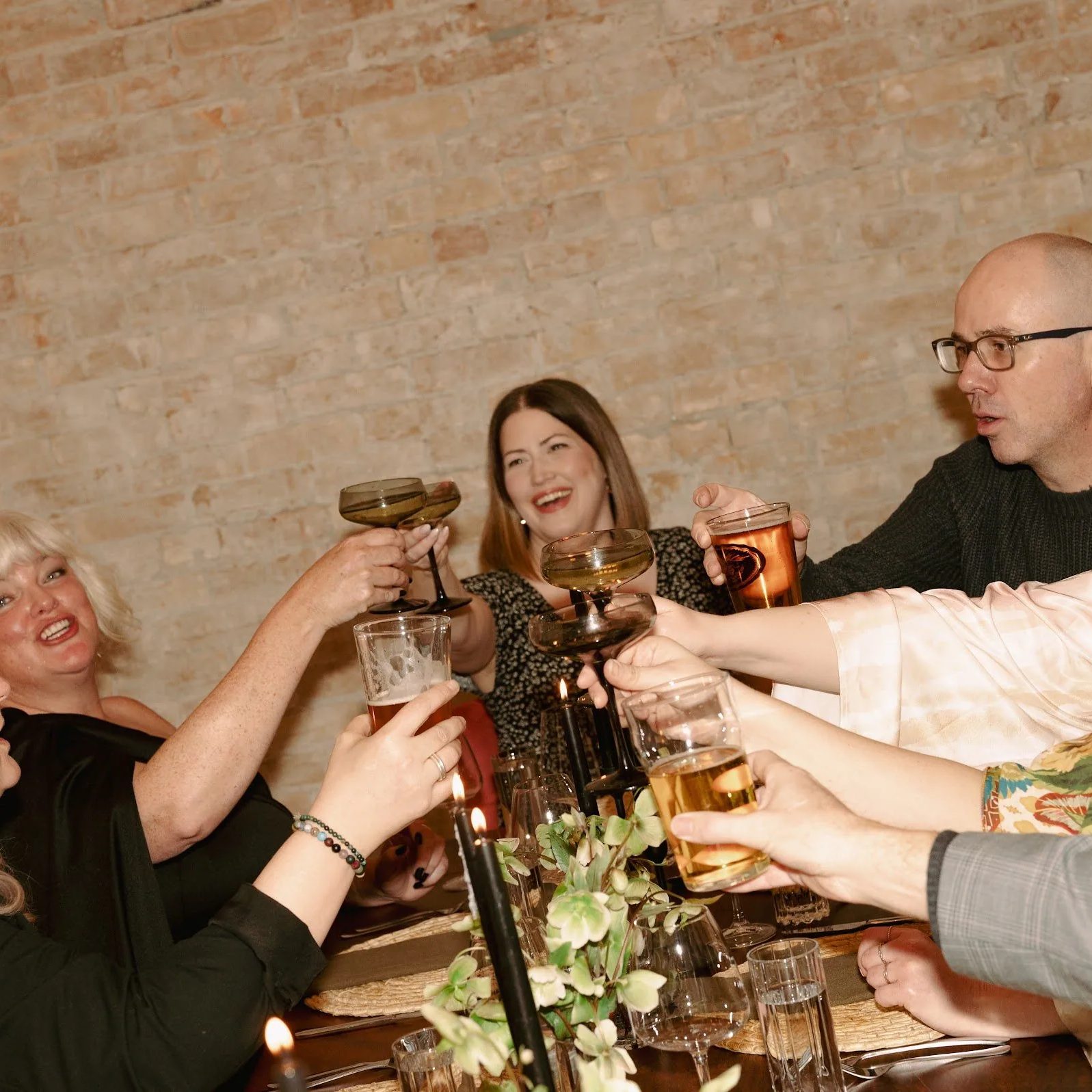 Guests raising a toast at a long candlelit table against the exposed brick wall at Den 1880