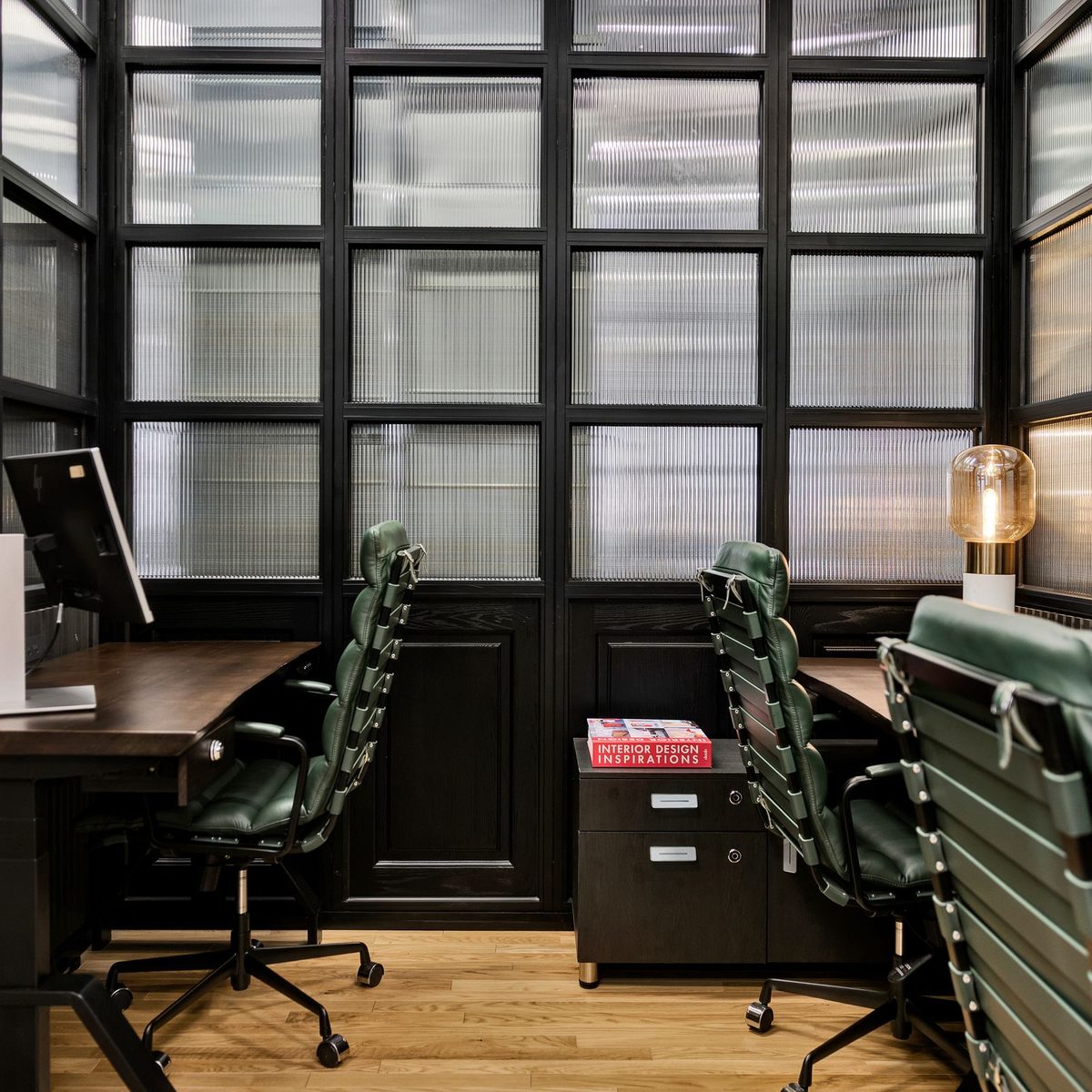 A furnished private office at Den 1880 with a monitor on the desk, a filing cabinet and green leather chairs