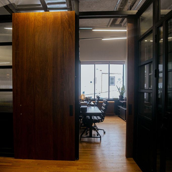 A sliding wood suite door standing open onto a private office with desks and a window at Den 1880