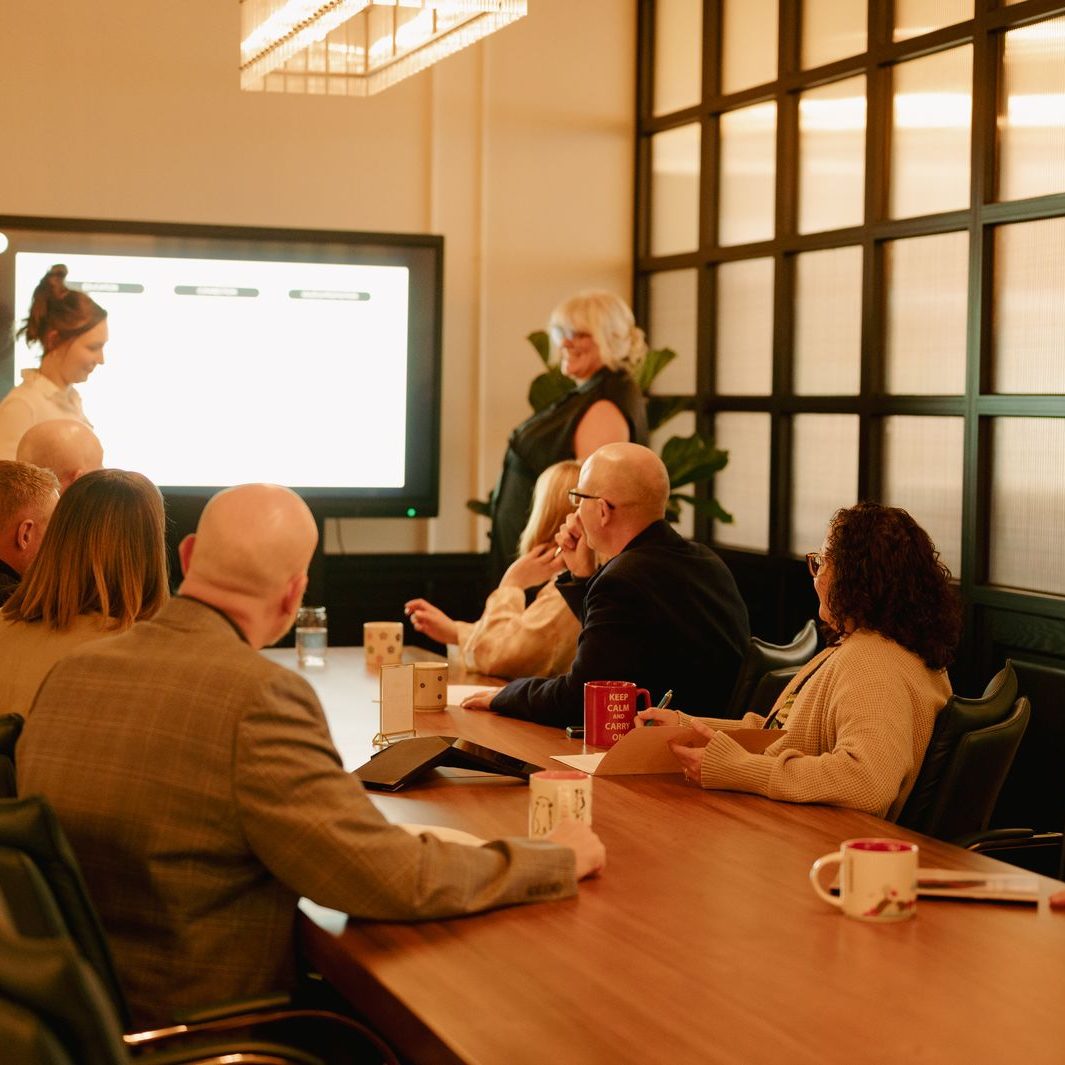 A presenter running a session on the wall screen for a team seated around the boardroom table at Den 1880
